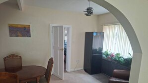 Uma área de refeições com mesa e cadeiras, uma geladeira e vista para uma varanda com plantas através de uma porta.
