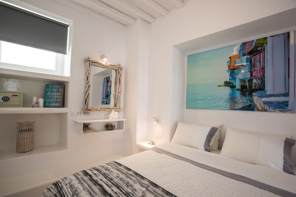 A bedroom with a bed, a painting of a coastal scene, a mirror, and shelves with items.