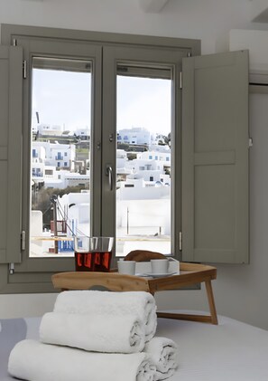 A room with a bed, a window with a view of white buildings, a wooden table with a bowl and a cup, and rolled towels on the bed. - Christy’s Three-Level Villa in Mykonos Town (Mykonos)