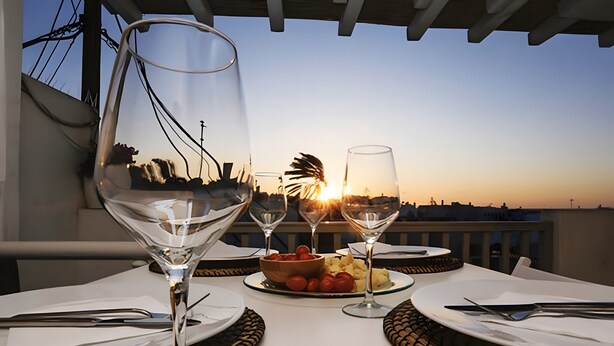 Ein Tisch für zwei Personen mit Weingläsern, Tellern und einer Schüssel Essen auf einem Balkon mit Blick auf eine Stadtlandschaft bei Sonnenuntergang.