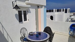 Un edificio blanco con balcón, una mesa redonda con sillas, mantel de tela azul y blanco, y una manguera azul.
