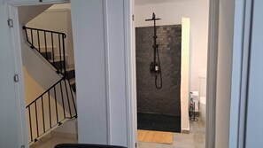 Una escalera con barandilla negra conduce a un cuarto con pared de ladrillo negro, ducha y retrete.