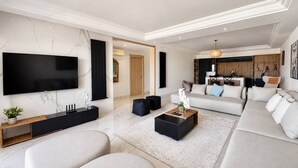 Un salon moderne avec un téléviseur à écran plat, un ensemble de canapés, une table basse et un coin cuisine à l’arrière-plan.