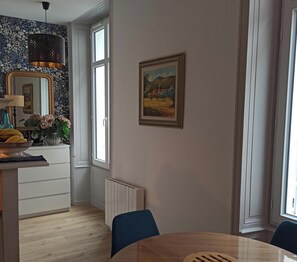 A dining area with a round table, chairs, a radiator, a window, and a painting on the wall. - Very pretty studio 100 meters from the main beach and pedestrian streets. (Les Sables-d'Olonne)
