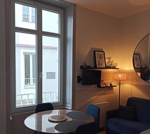 A room with a round table, a blue sofa, a floor lamp, a TV, and a large window with a view of a building. - Very pretty studio 100 meters from the main beach and pedestrian streets. (Les Sables-d'Olonne)