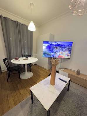 Ein modernes Wohnzimmer mit einem runden Esstisch, einem an der Wand montierten Fernseher und einer Vase mit Trockenblumen auf einem Couchtisch.