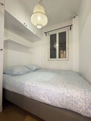 Ein Einzelbett mit gemustertem Bettbezug, eine Hängeleuchte und ein Fenster mit Blick auf einen Raum.