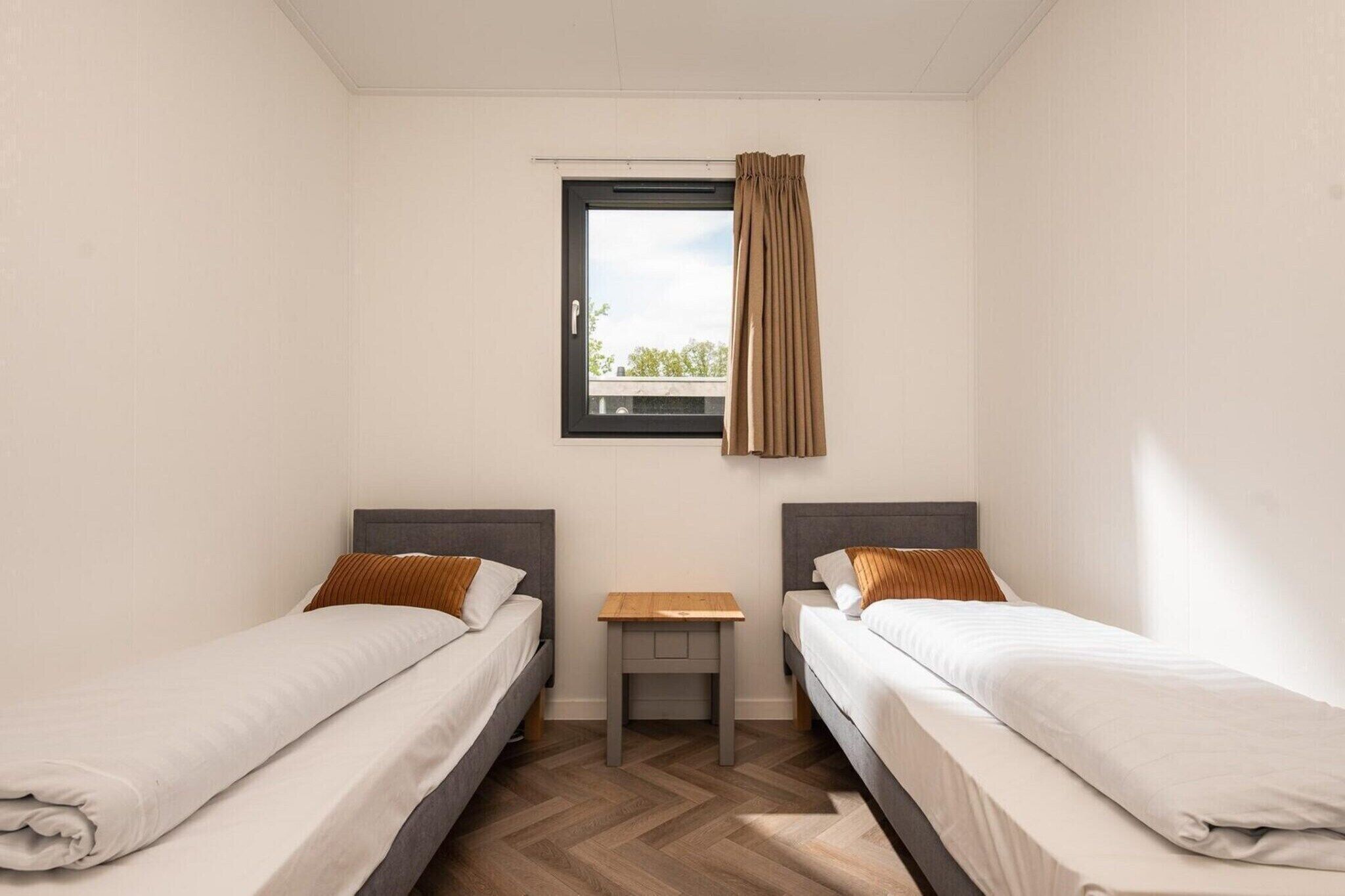 A twin room with two beds, a small table, a window with curtains, and a view of greenery outside.