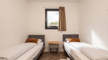 A twin room with two beds, a small table, a window with curtains, and a view of greenery outside.