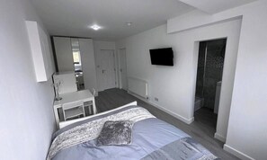 A bedroom with a bed, a desk, a chair, a TV, and a shower area.