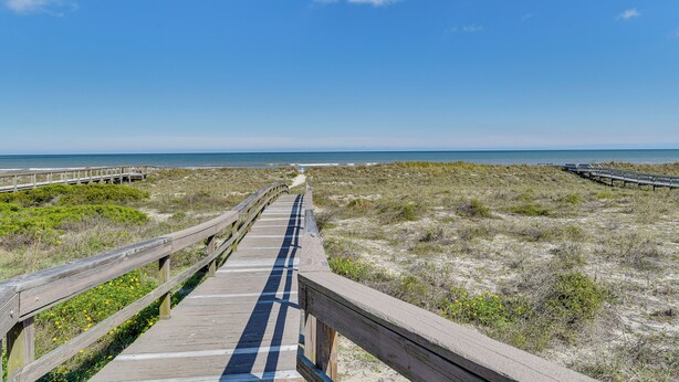 Ein hölzerner Steg führt zu einem Strand mit Dünen und blauem Himmel.