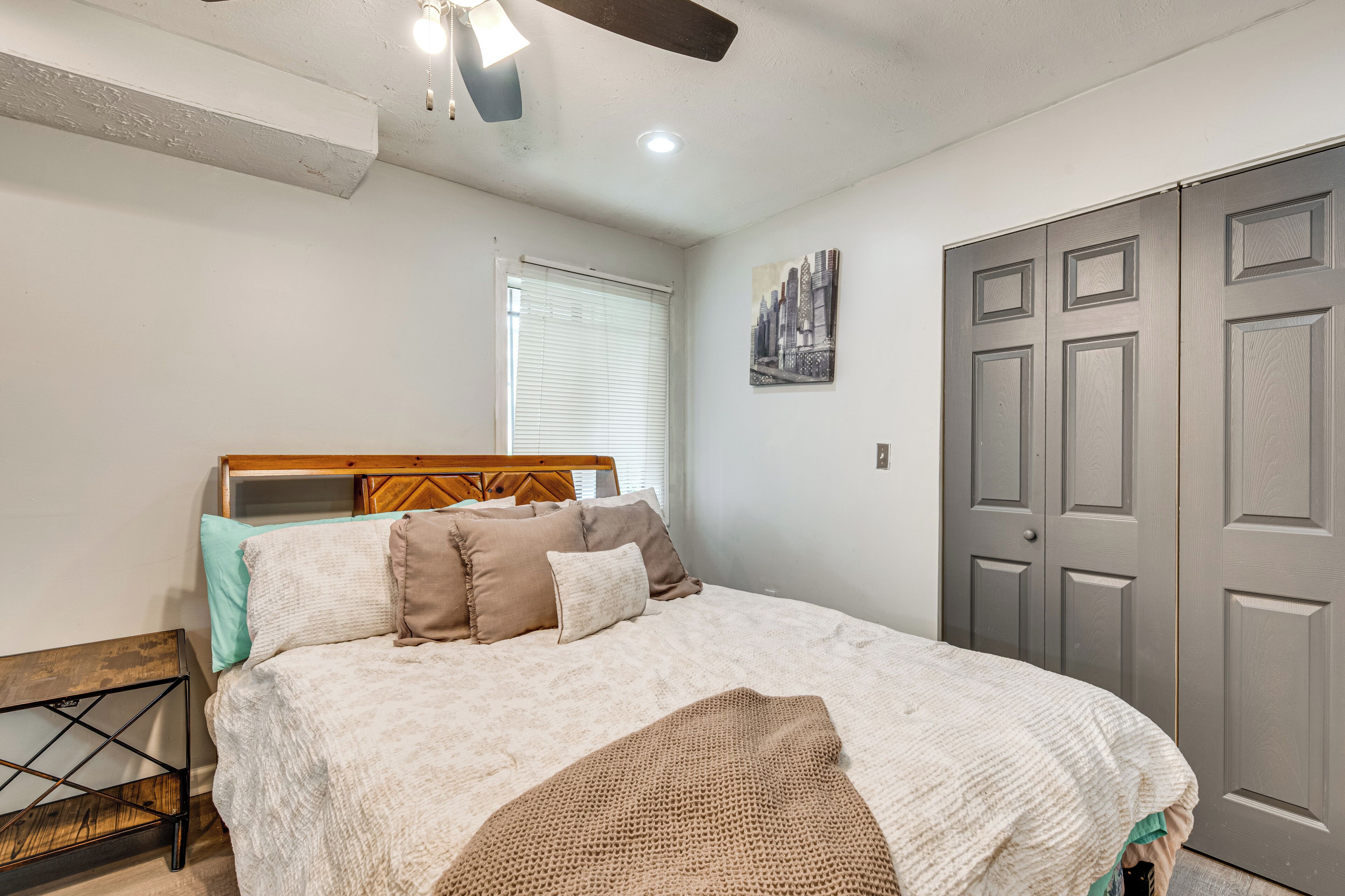 Un dormitorio con cama, cabecera de madera, ventana con persianas, puerta gris y un cuadro de paisaje urbano en la pared.