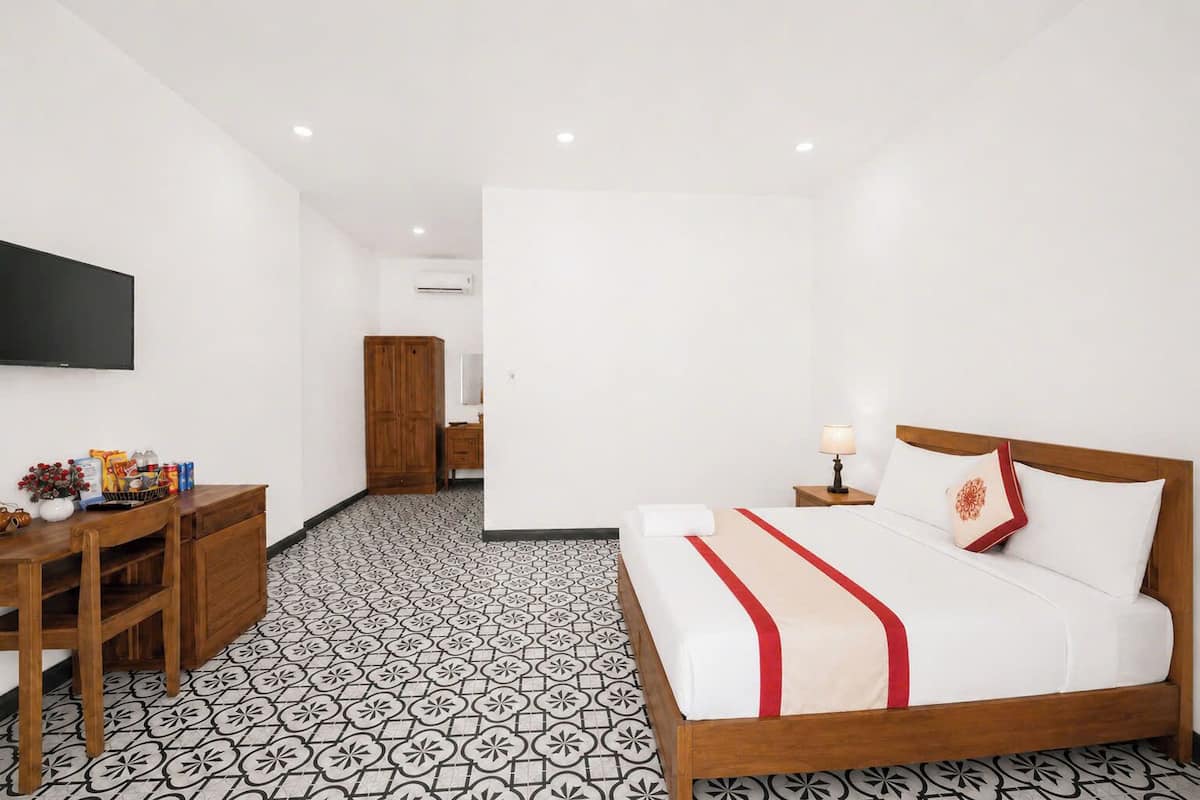A hotel room with a bed, bedside table, lamp, TV, wardrobe, and patterned floor.