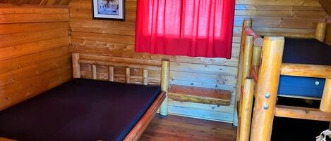 A wooden cabin with a bunk bed, a single bed, a red curtain, and a framed picture on the wall.