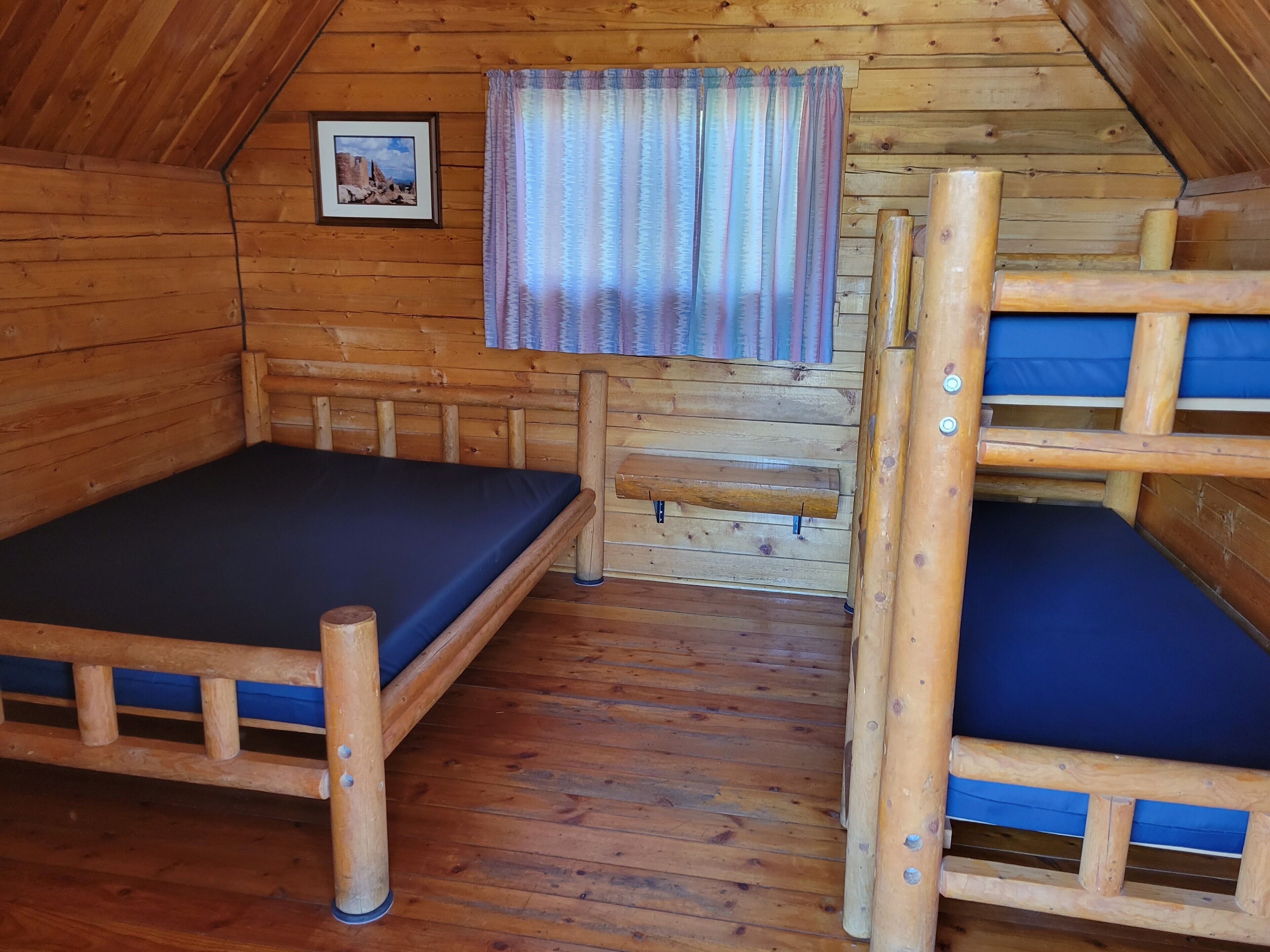 A wooden cabin with bunk beds, a window with curtains, and a framed picture on the wall.