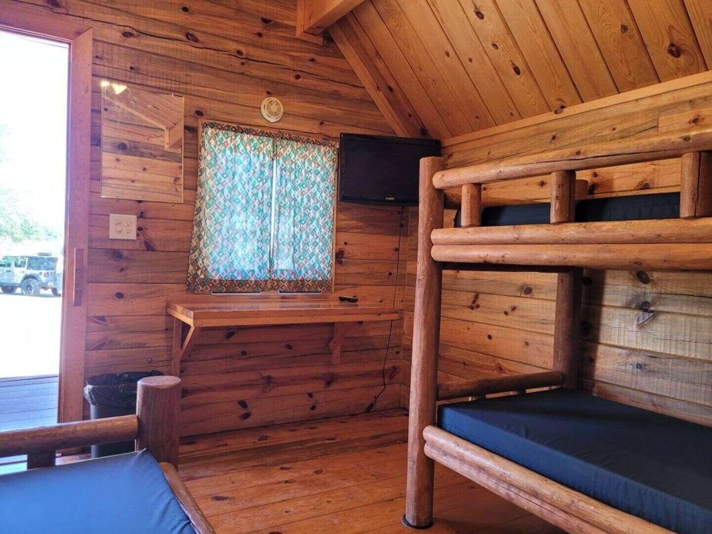 L’intérieur d’un chalet en bois, avec un lit superposé, une fenêtre ornée de rideaux à motifs et un téléviseur fixé au mur.