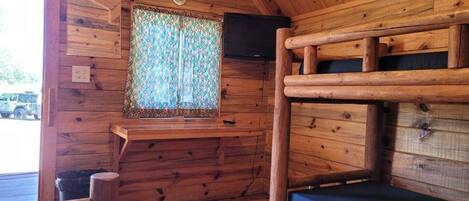 L’intérieur d’un chalet en bois, avec un lit superposé, une fenêtre ornée de rideaux à motifs et un téléviseur fixé au mur.