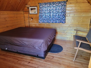 A wooden cabin bedroom with a bed, a chair, a lamp, and a patterned curtain. - Cortez RV Resort (Cortez)