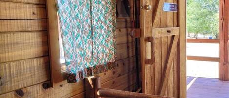 Interior de una cabaña de madera con una cama, un cortinaje estampado y una puerta que da al exterior.