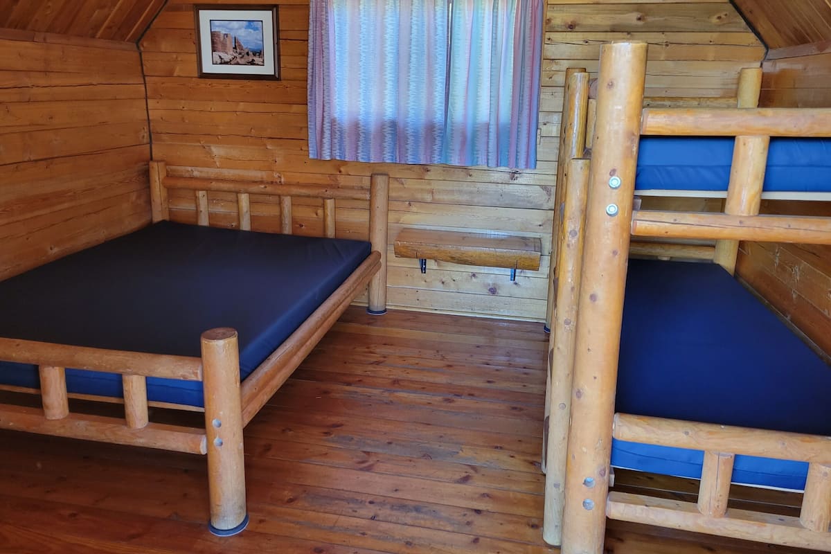 A wooden cabin room with bunk beds, a small window with curtains, and a framed picture on the wall.