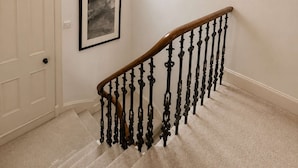 Eine Treppe mit hölzernem Geländer und schwarzen Spindeln, eine weiße Tür, ein gerahmtes Bild an der Wand und eine Pendelleuchte.