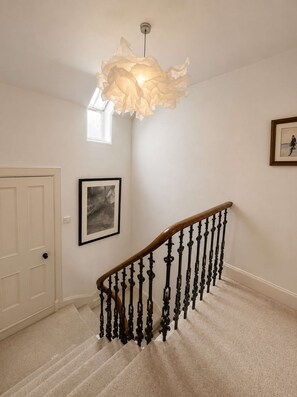 Un escalier avec une rampe en bois et des balustres noires, une porte blanche, un tableau encadré au mur et un luminaire suspendu.