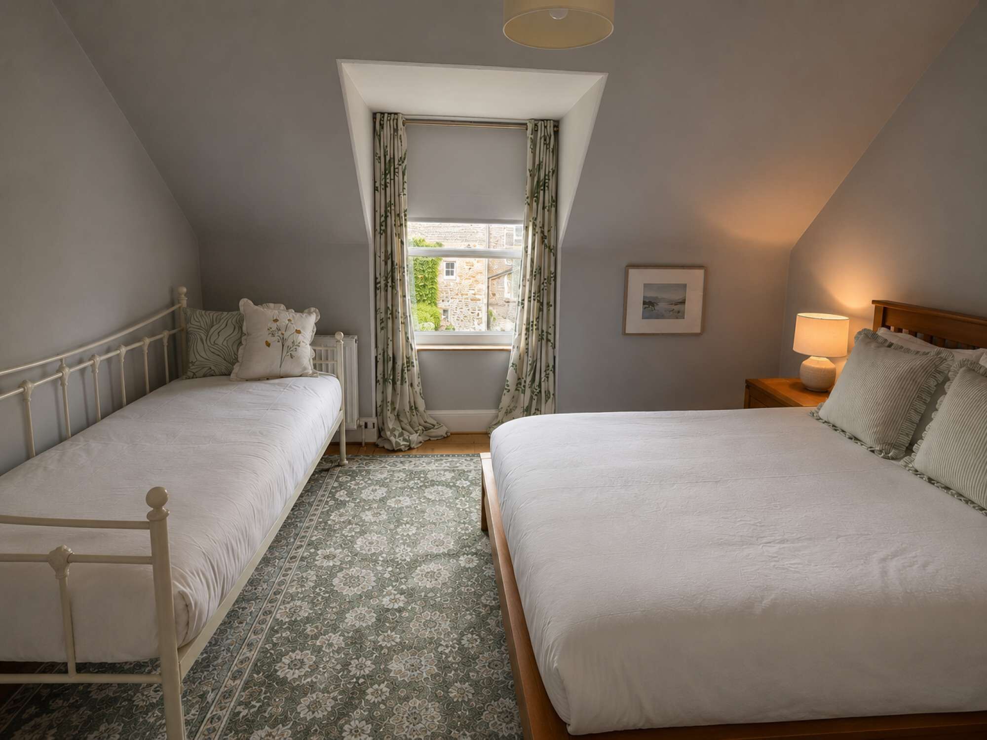 Een slaapkamer met twee bedden, een raam met gordijnen, een bedlampje en een lijst met een afbeelding aan de muur.
