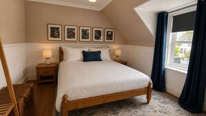 Ein Schlafzimmer mit einem hölzernen Bett, zwei Nachttischen mit Lampen, einer Kommode, einem Fenster mit Vorhängen und vier gerahmten Bildern an der Wand.