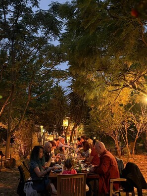 Un espace extĂ©rieur amĂ©nagĂ© pour les repas, avec des tables et des chaises, entourĂ© dâarbres et avec un bĂątiment en arriĂšre-plan.