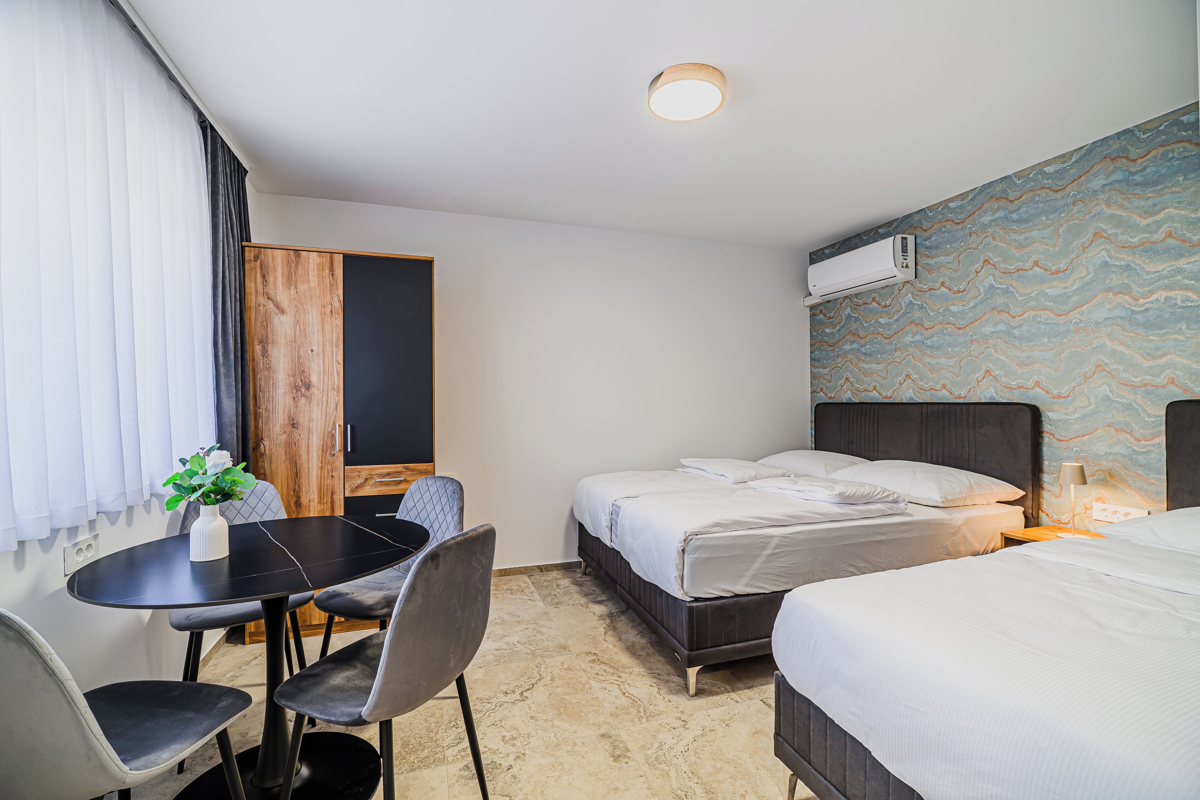A hotel room with two beds, a dining table with chairs, a small plant, and a wall-mounted air conditioner.