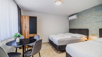 A hotel room with two beds, a dining table with chairs, a small plant, and a wall-mounted air conditioner.