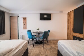 Habitación de hotel con dos camas, un televisor montado en la pared, una mesita redonda con sillas y un armario de madera.