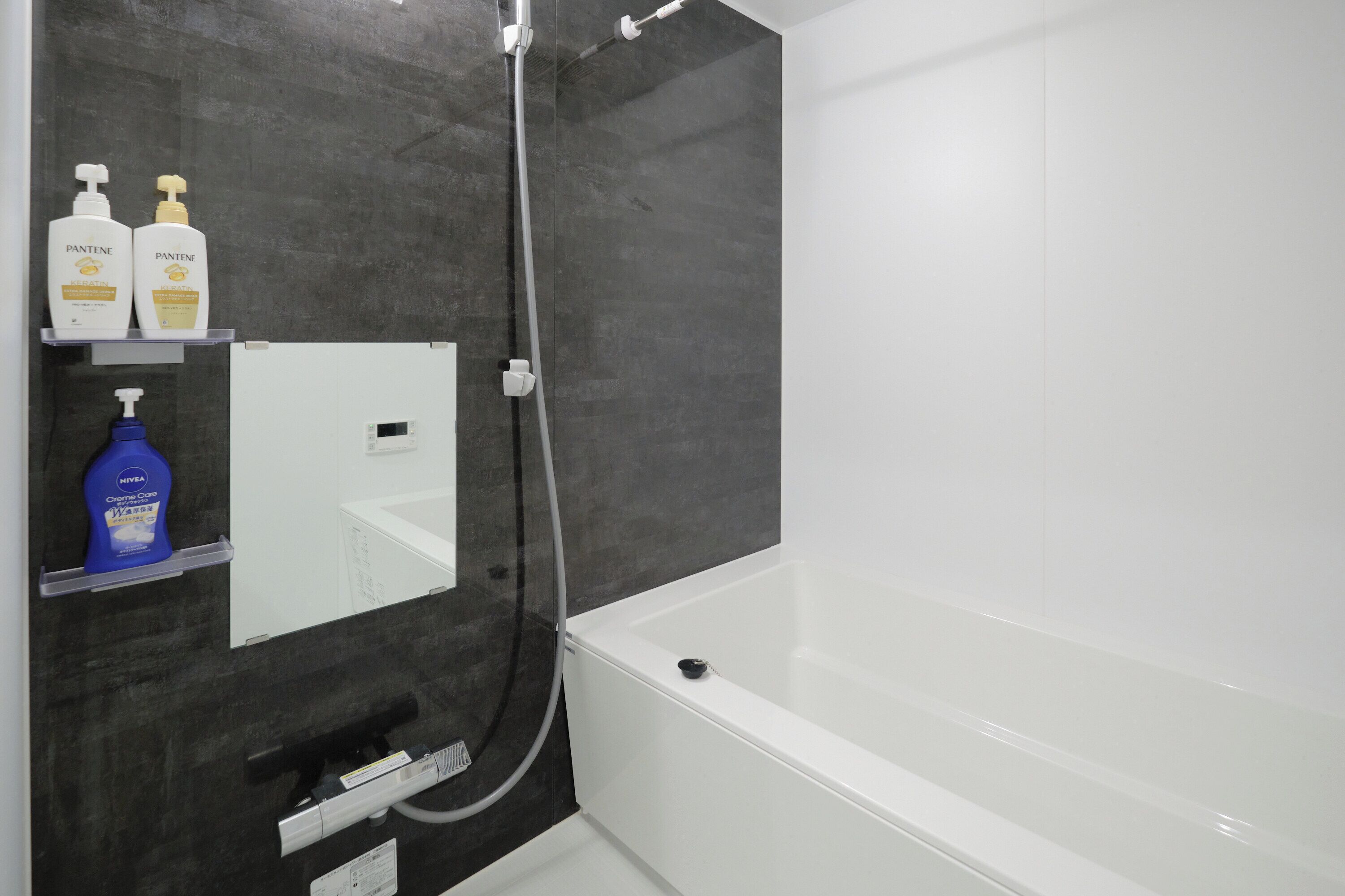A bathroom with a bathtub, a shower, and a mirror. There are two bottles of Pantene shampoo and a bottle of Hema cream on the shelf.