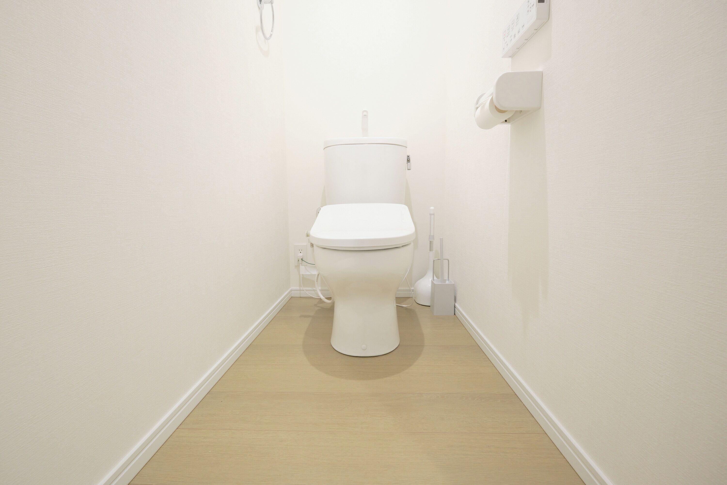 A small, white toilet in a narrow, white-walled bathroom with a wooden floor.