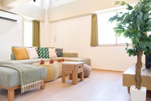 A modern living room with a sofa, two armchairs, a coffee table, and a small wooden stool. There is a potted plant on a table and a window with curtains. - Liverty Place Nakano 2 (Tokyo)