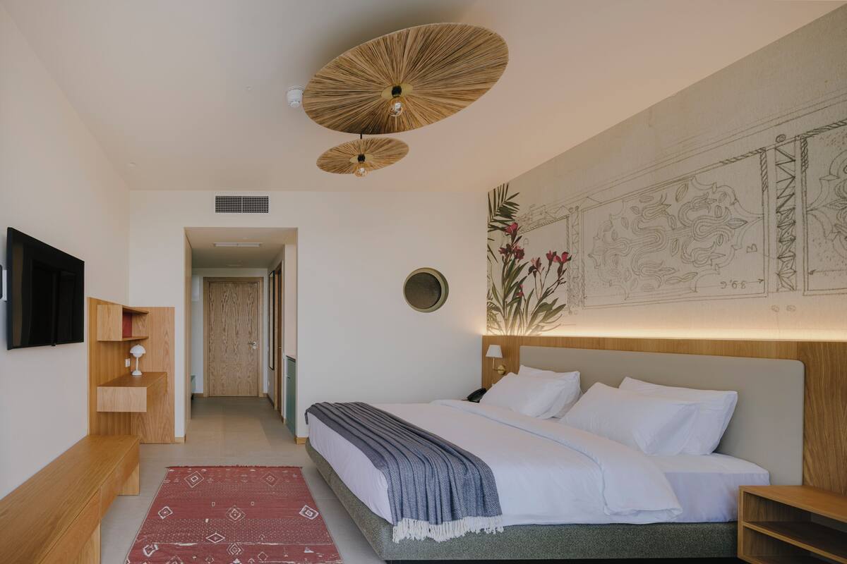 A bedroom with a bed, a wooden bench, a red patterned rug, a wall-mounted TV, and a ceiling fan.
