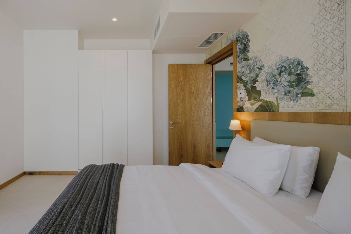 A hotel room with a bed, a wooden door, a wall-mounted lamp, and a floral wallpaper.