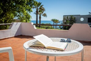 An open book on a white table with a pair of glasses beside it, set on a terrace with a view of the sea and palm trees. - GuestHost - Lovely Sea View Villa with Parking (Porto Columbu-Perd'È Sali)