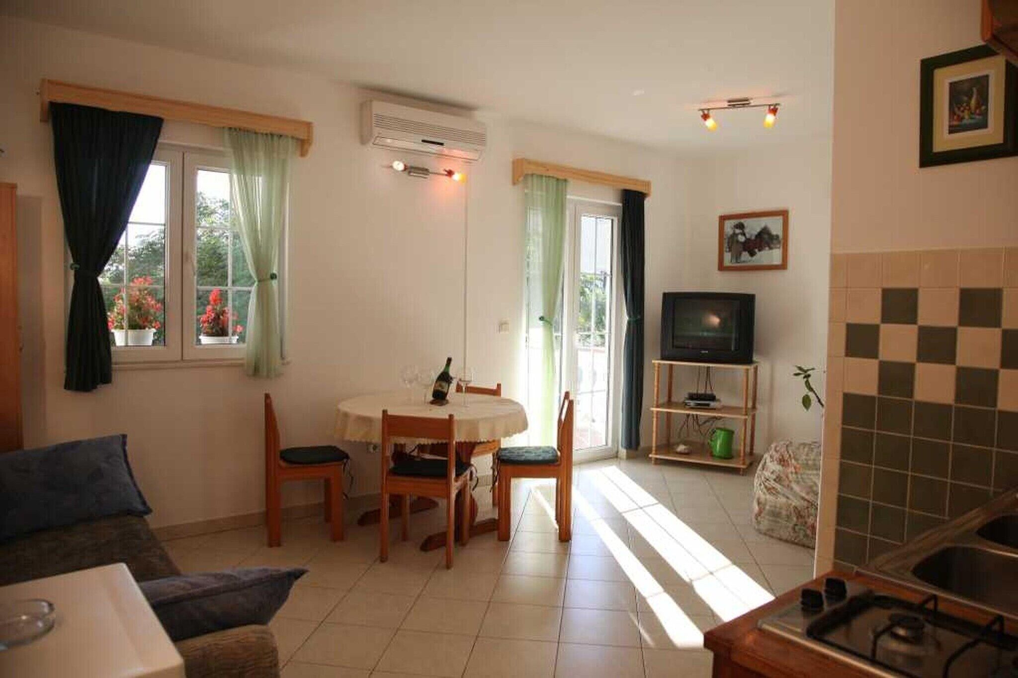 Ein Wohnzimmer mit einer Couch, einem Esstisch mit Stühlen, einem Fernseher und einer Küchenzeile inklusive Spüle und Herd.