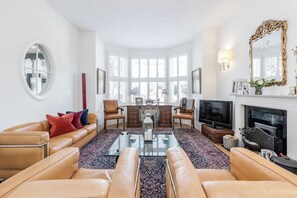 Uma sala de estar espaçosa com sofás de couro, uma mesa de centro de vidro, lareira, televisão e uma grande janela com vista para edifícios.