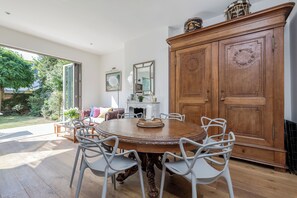 Ein Essbereich mit einem runden Holztisch, umgeben von weißen Stühlen, einem Holzschrank mit dekorativen Schnitzereien und Blick ins Freie durch große Glasüren.