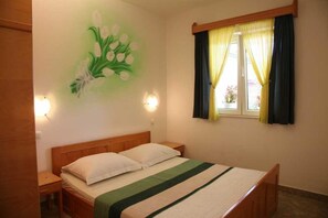 Ein Schlafzimmer mit einem Bett, einem Nachttisch, einer Wandlampe, einem Fenster mit Vorhängen und einem floralen Wandbild.