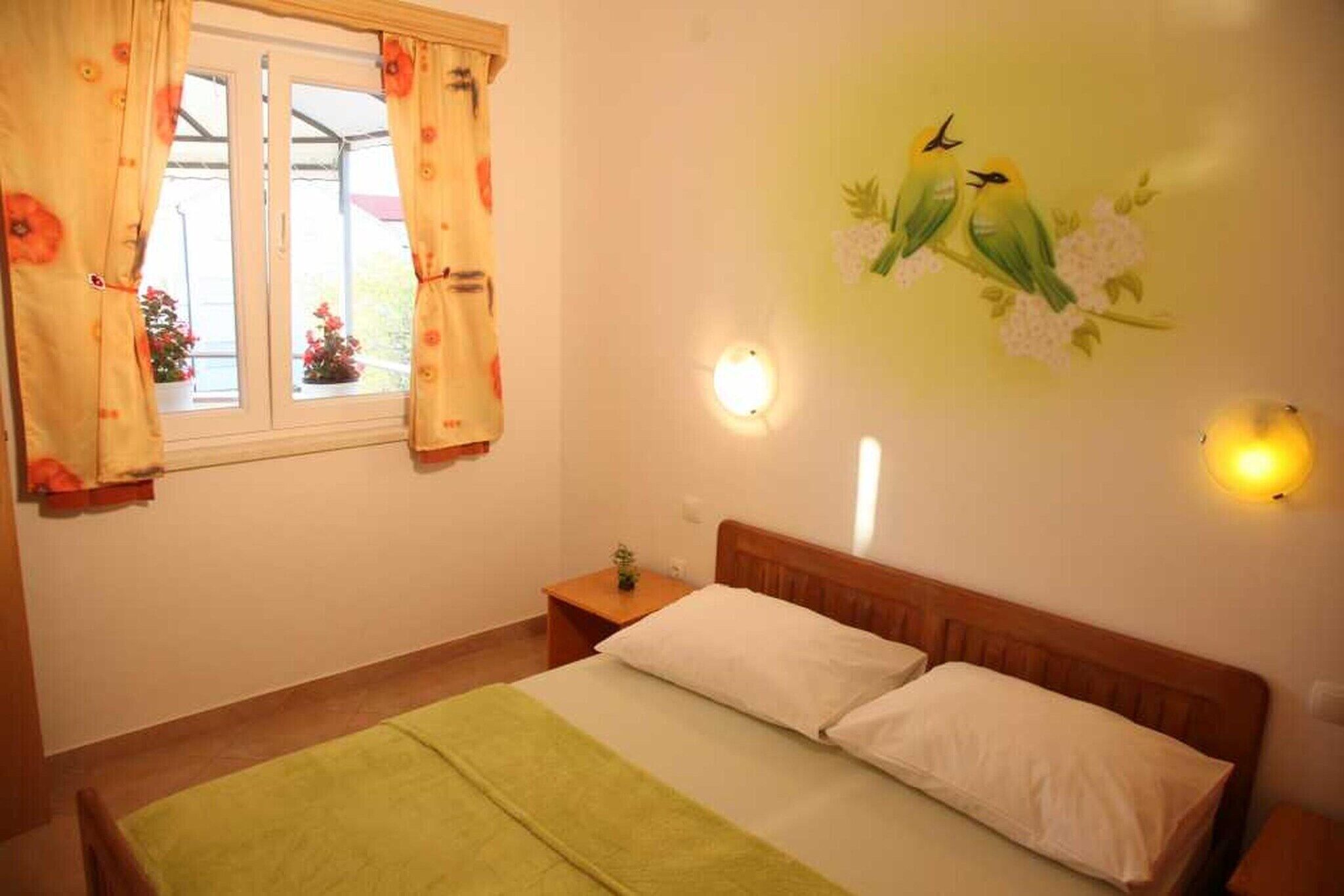 Ein Schlafzimmer mit Bett, Nachttisch, einem Fenster mit Vorhängen, Wandleuchten und einer Vogel-Wanddekoration.