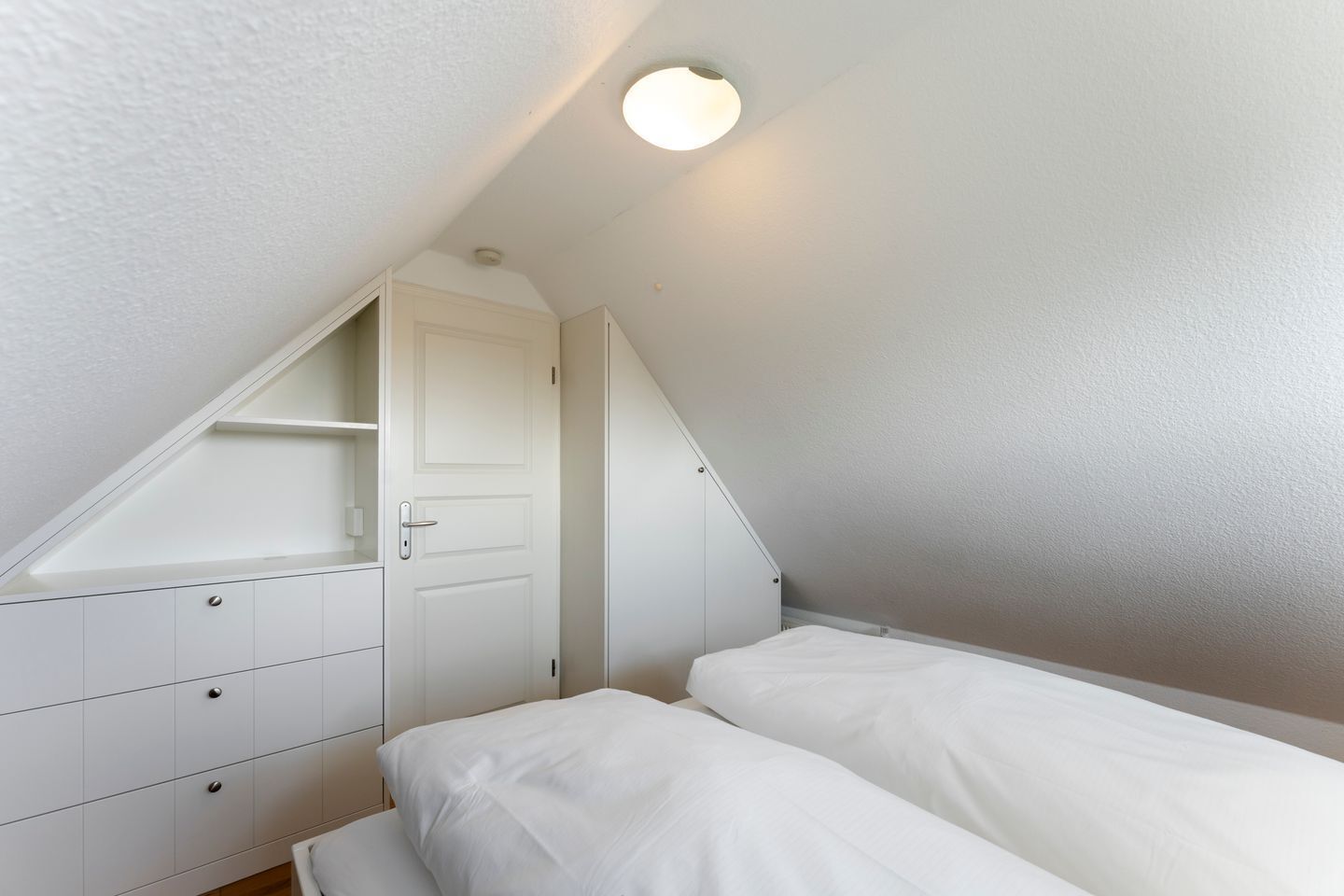 Une chambre avec un plafond en pente, un lit simple, une armoire blanche et une porte.