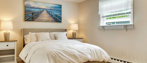 A bedroom with a bed, two bedside tables with lamps, a painting of a pier, and a window with blinds.