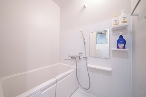 Uma casa de banho branca com banheira, chuveiro e espelho. Existem prateleiras com produtos de higiene pessoal.