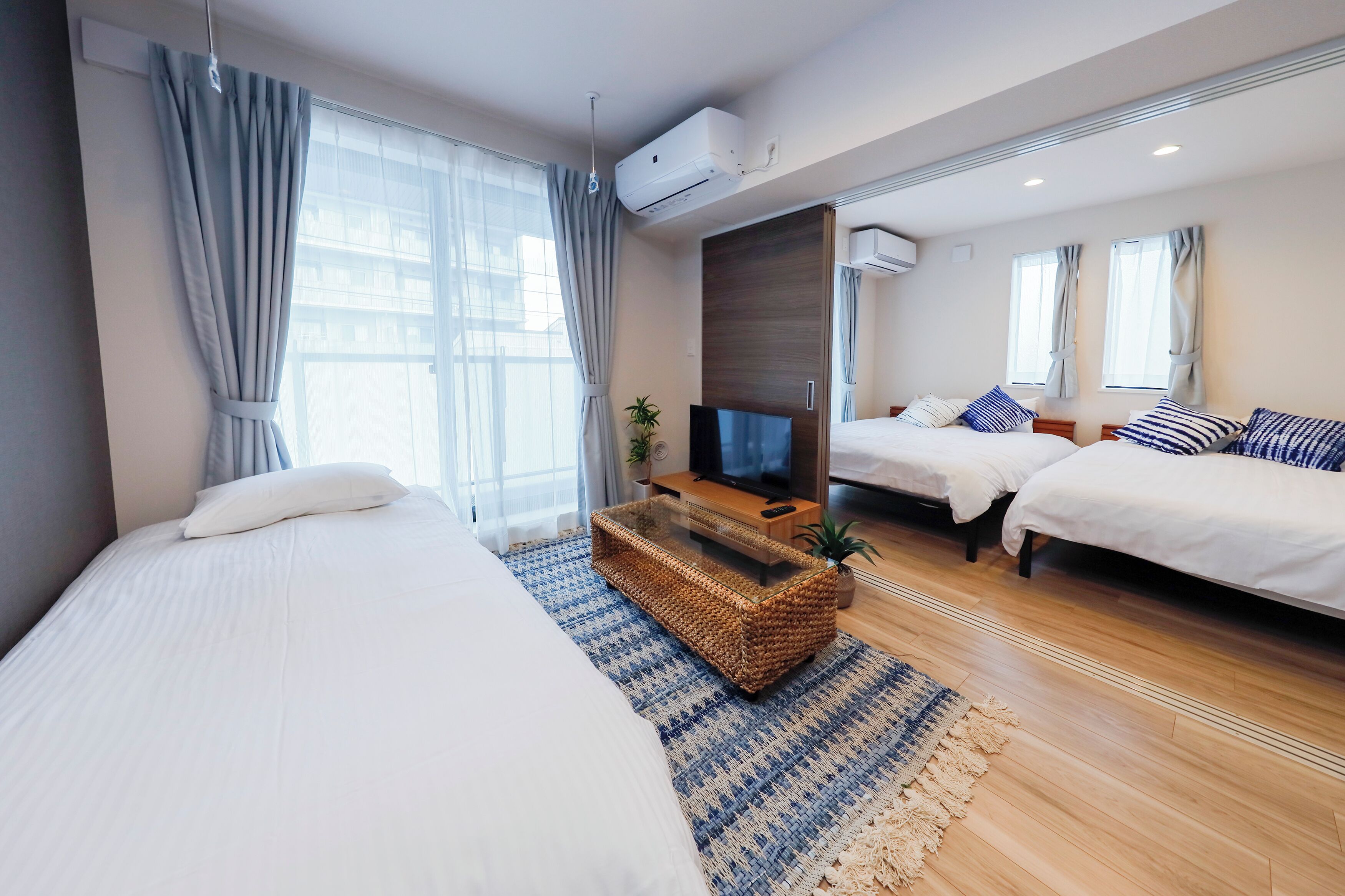 Quarto de hotel com duas camas, televisão, mesa de centro de fibra vegetal e uma janela grande com cortinas.