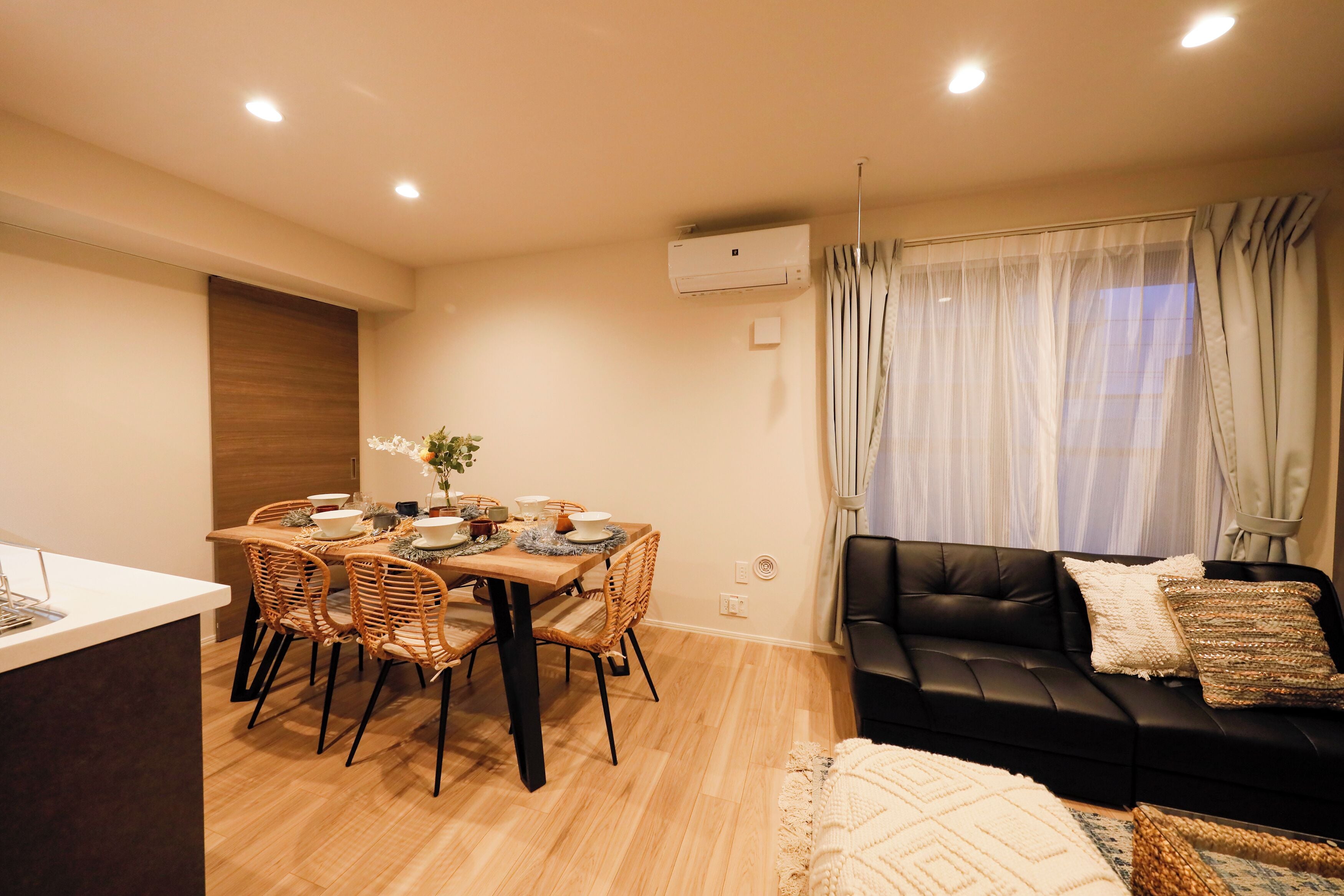 Una sala moderna con un sofá de cuero negro, una mesa de comedor con sillas de mimbre y un ventanal con cortinas traslúcidas.