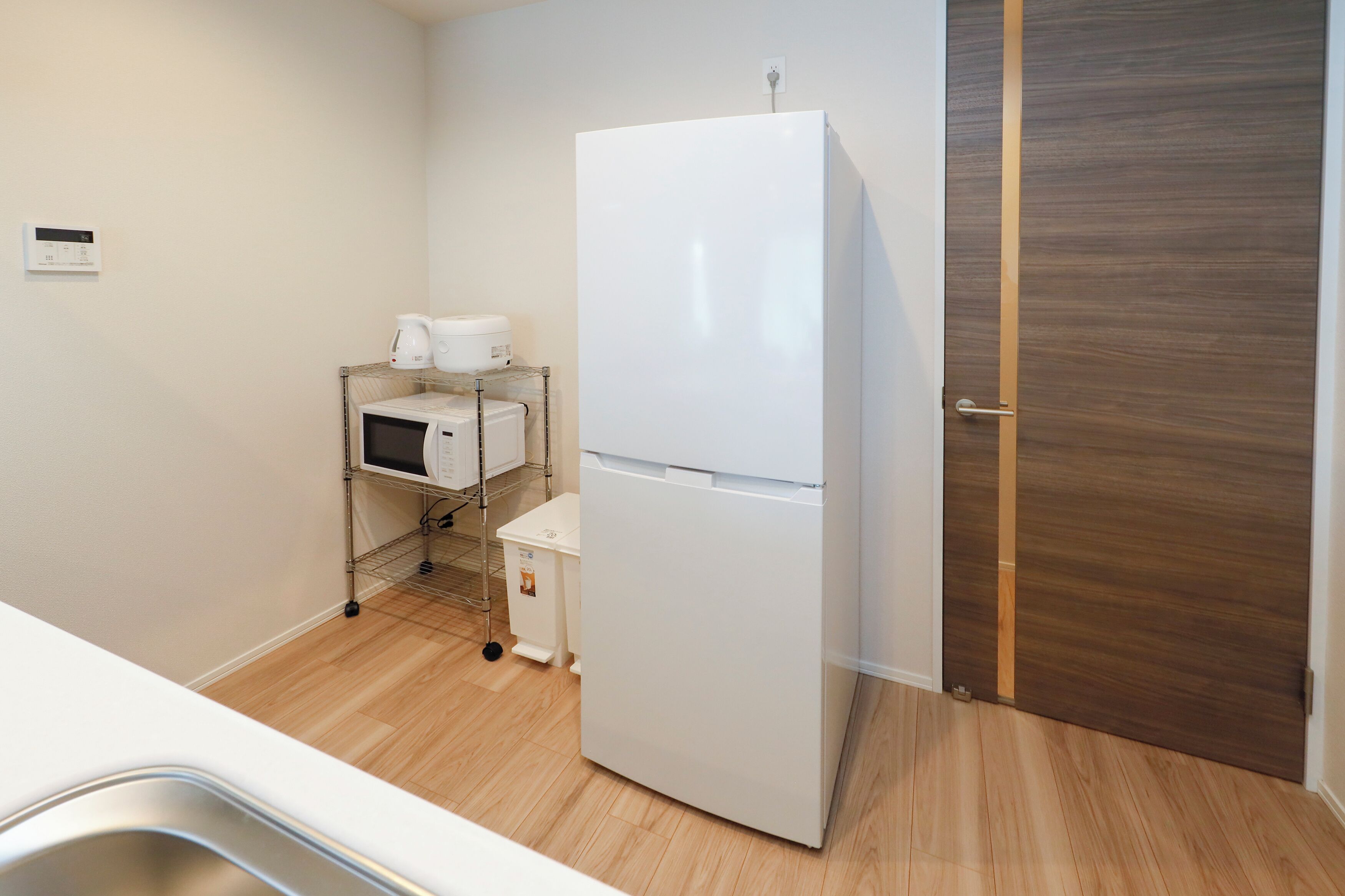 Una cocina compacta con un refrigerador blanco, microondas y cafetera sobre una estantería metálica, una puerta de madera y un lavamanos de acero inoxidable.