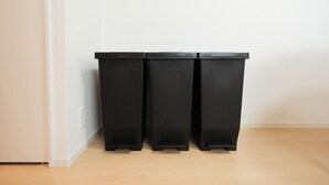 Tres contenedores de basura negros alineados contra una pared blanca con suelo de madera.